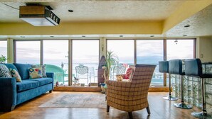 A spacious living room with large windows, a blue sofa, a wicker chair, and a bar area.