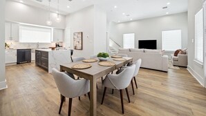 A modern kitchen with white cabinets, a dining table with chairs, and a living area with a sofa and TV. - Luxury Group Retreat | Rooftop Deck + Chef's Kitchen | Sleeps 10 (Houston)