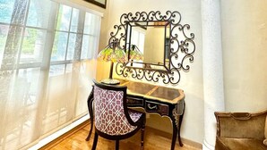 Une pièce comprenant un bureau, une chaise et un grand miroir. Les murs sont ornés de tableaux encadrés et il y a une fenêtre avec des stores.