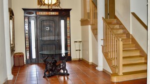 Un hall d’entrée avec un escalier en bois, une table en verre surmontée d’une sculpture métallique décorative et une porte en bois foncé agrémentée d’un panneau décoratif.