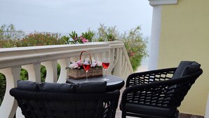 A balcony with two wicker chairs and a table, a wicker basket with flowers and wine glasses, and a view of the sea.