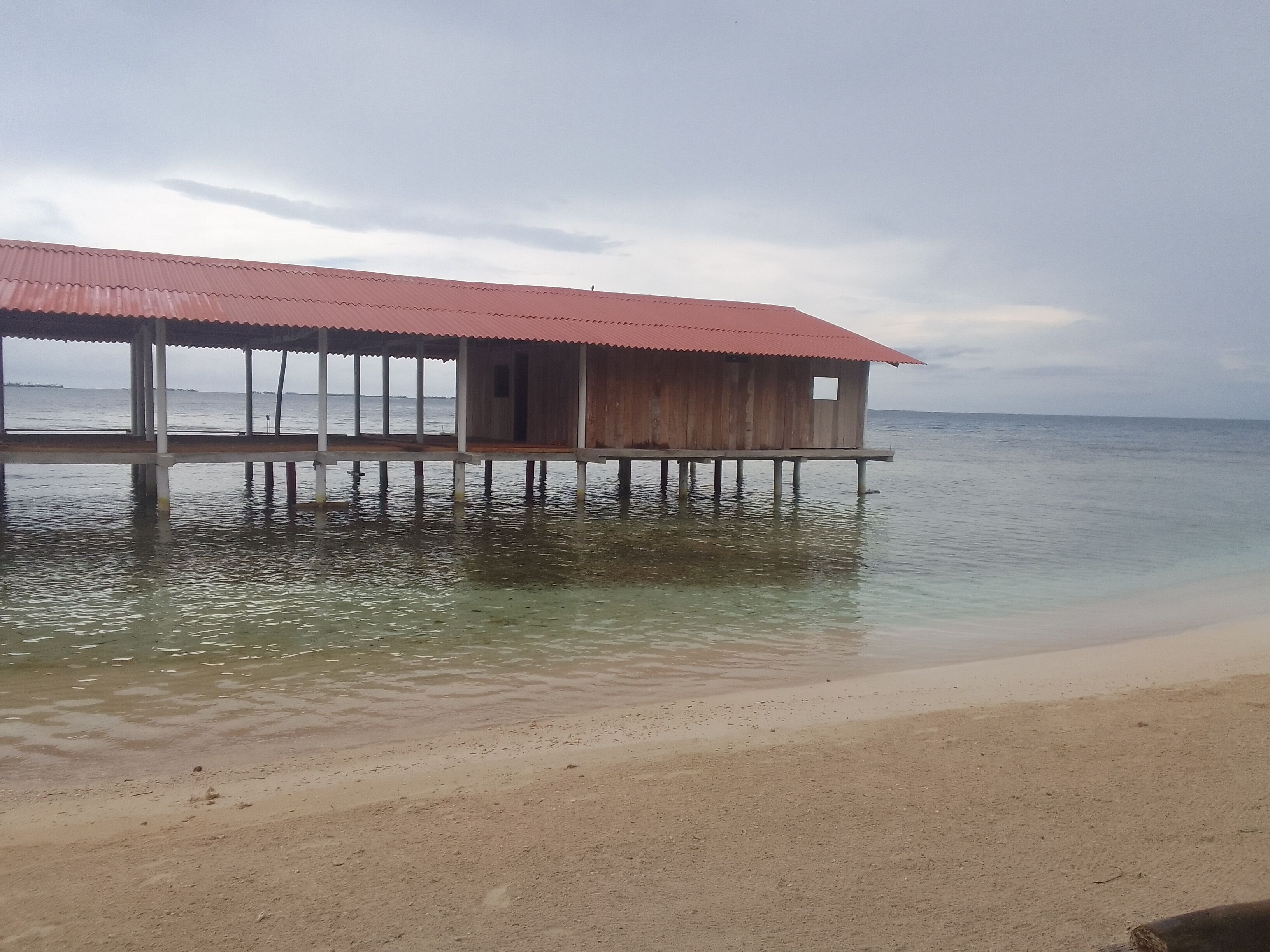 A wooden structure with a red roof on stilts over water, adjacent to a sandy beach.