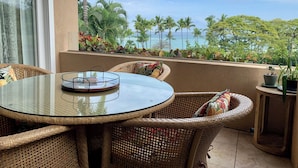 Une table ronde en verre, des chaises en osier, une petite table d’appoint et une vue sur l’océan, avec des palmiers en arrière-plan.