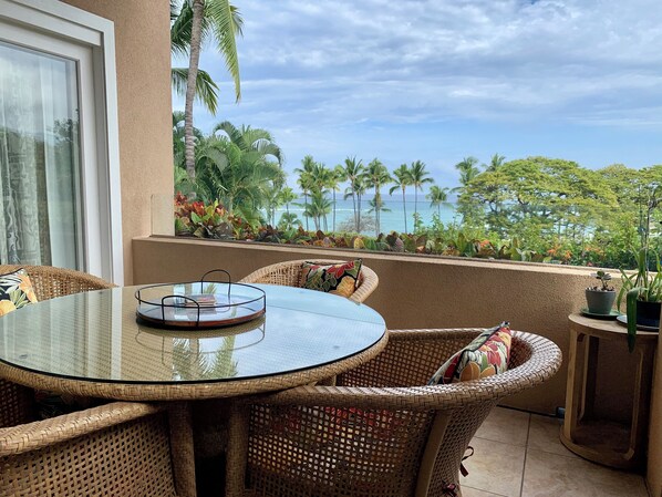 A round glass table with wicker chairs, a small side table, and a view of the ocean with palm trees.