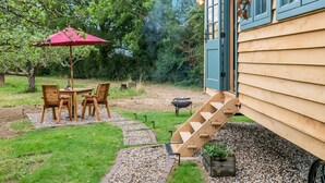 A wooden house with a blue exterior, a wooden deck, a red umbrella, and a stone pathway. - Set among the fruit trees with views of the fields. (Ongar)