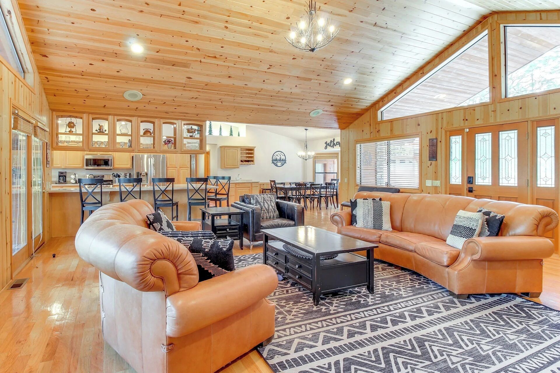 A spacious living area with leather sofas, a coffee table, and a dining table with chairs. The room has wooden walls and ceiling, and large windows.