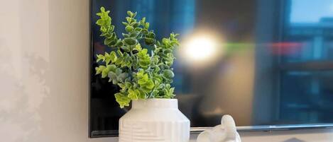Un florero blanco con plantas verdes sobre una consola de madera junto a una pared blanca con un amplio ventanal.