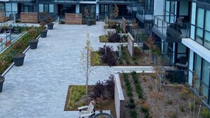 A modern multi-story building with large windows and balconies, a courtyard with seating and planters, and a clear sky. - Amazing 2BR/2BA Resort Condo w/ Balcony & Parking! (Innisfil)