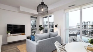 A modern living room with a grey sofa, a flat-screen TV, a dining table, and large windows offering a city view. - Amazing 2BR/2BA Resort Condo w/ Balcony & Parking! (Innisfil)