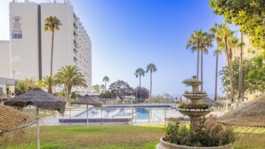 Zona de piscina con una fuente, rodeada de palmeras y un césped.