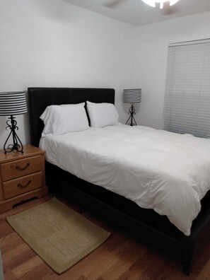 Un dormitorio con cabecera negra, ropa de cama blanca, una mesita de noche de madera con cajones y dos lámparas de noche.