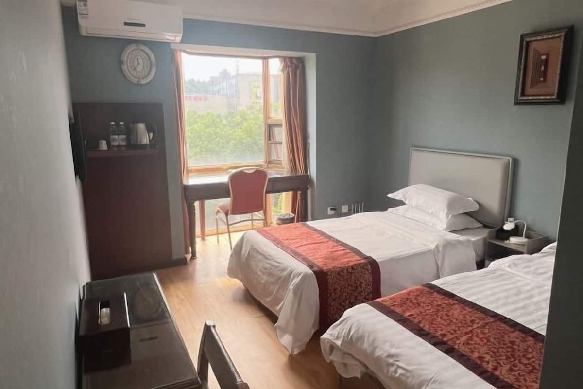 A hotel room with two beds, a desk, a chair, a window with curtains, and a clock on the wall.