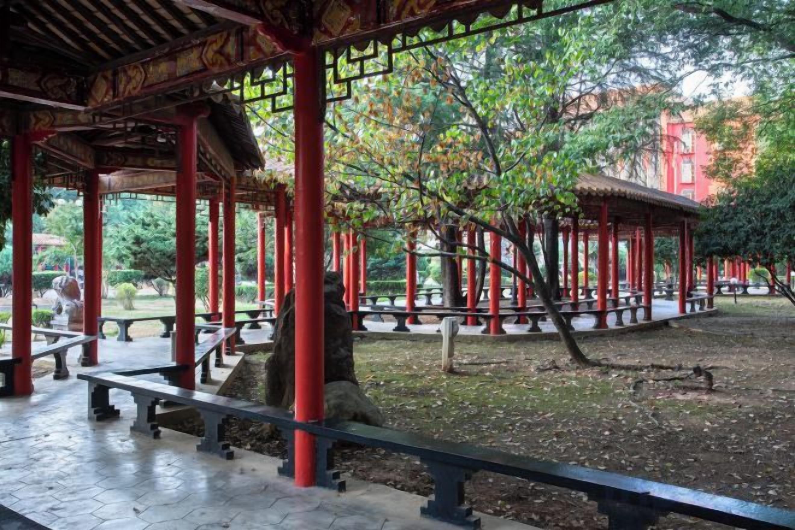 Um pavilhão com bancos e piso de azulejos, rodeado por árvores e um edifício vermelho ao fundo.