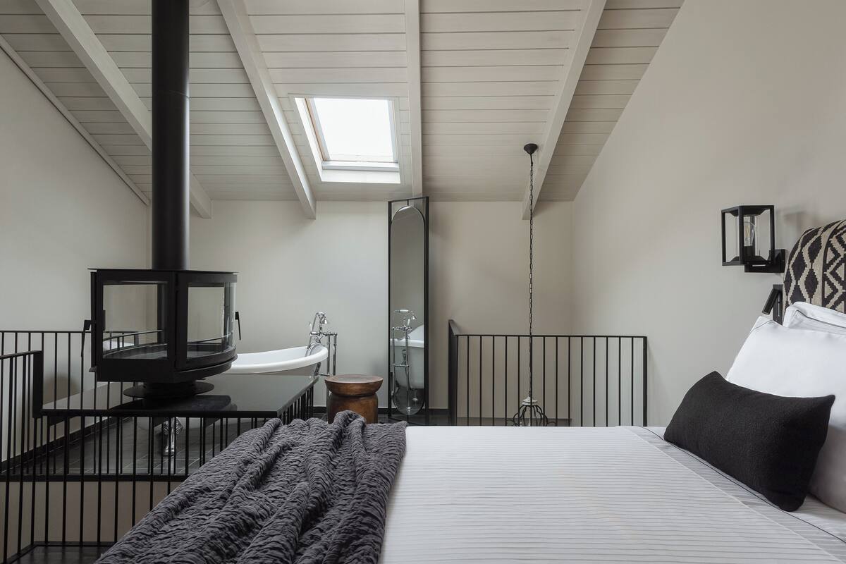 A bedroom with a bed, a black heater, a skylight, a bathroom with a sink and mirror, and a small wooden stool.