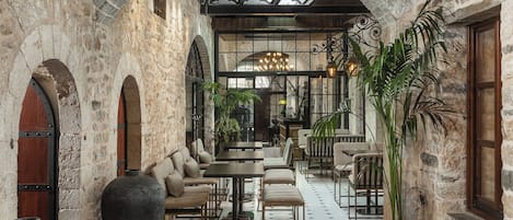 Un interior con paredes de piedra, piso de baldosas y un espacio de estar con mesas y sillas.