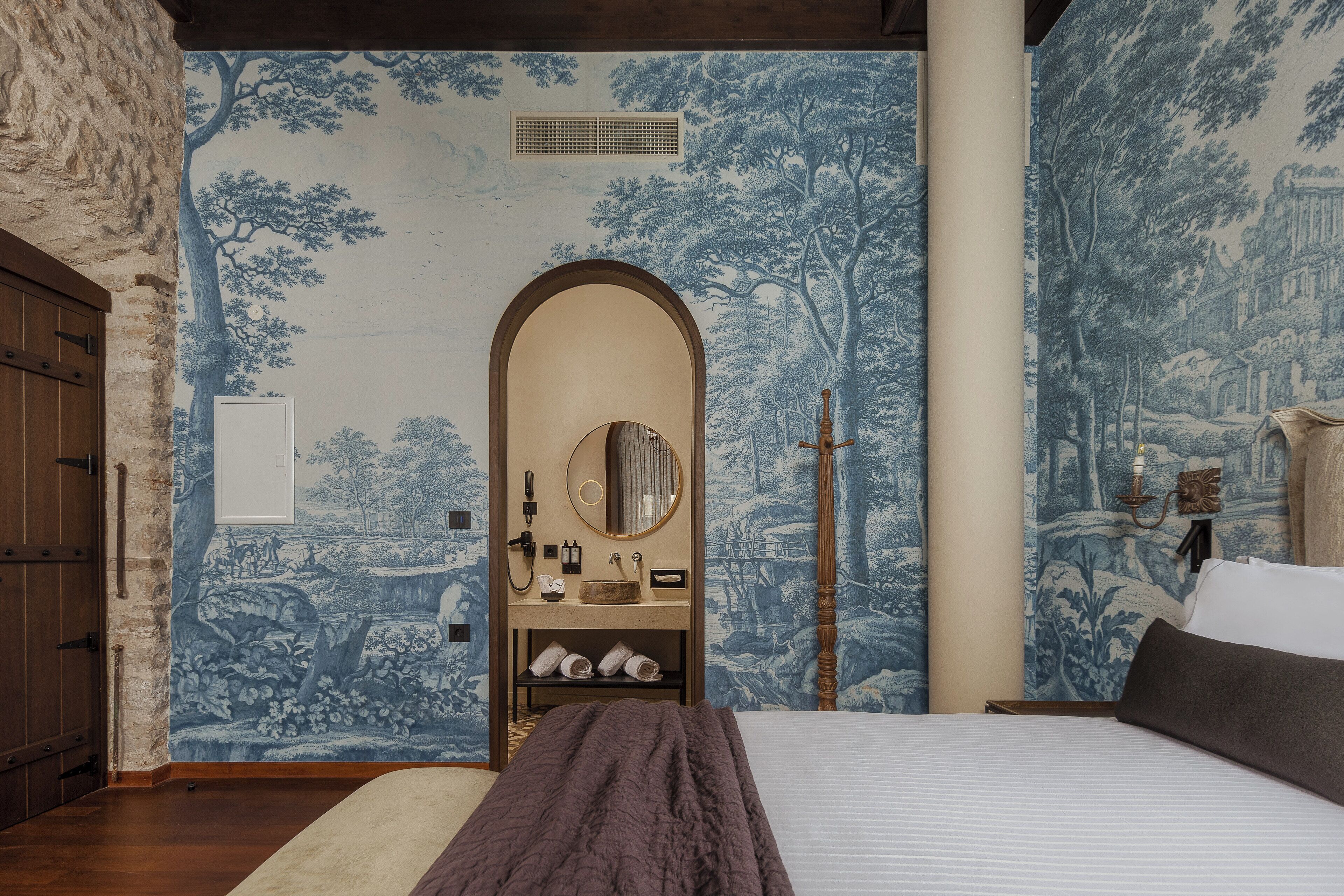 A bedroom with a large bed, a wooden wardrobe, a mirror, and a vanity with toiletries.