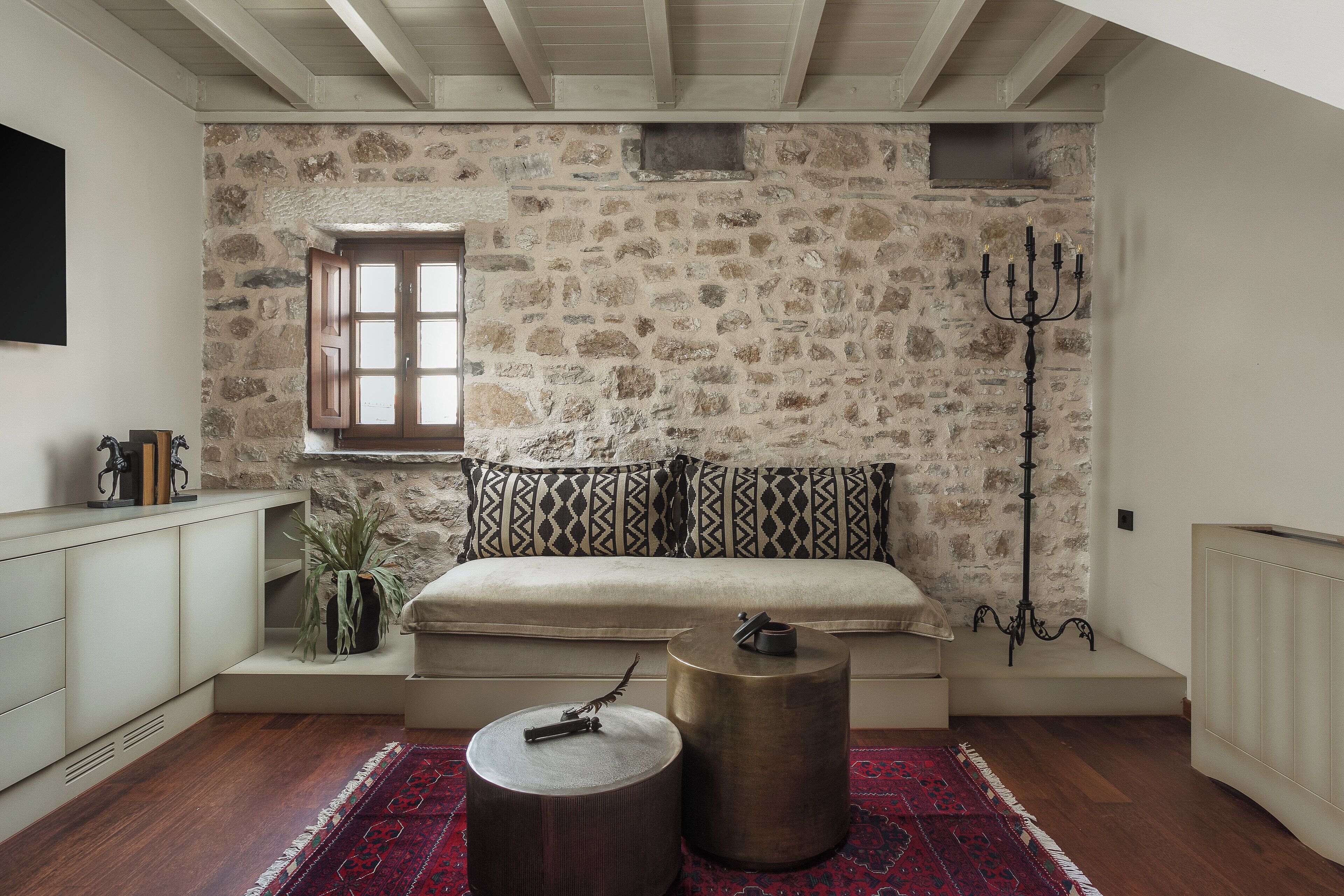 A cozy room with a stone wall, a cushioned bench, a round wooden table, and a red patterned rug.