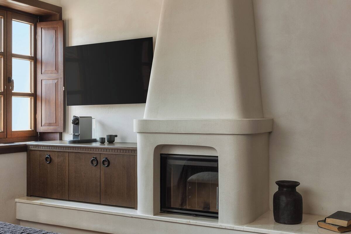 A bedroom with a bed, a wooden cabinet, a flat-screen TV, a white fireplace, and a window with wooden shutters.