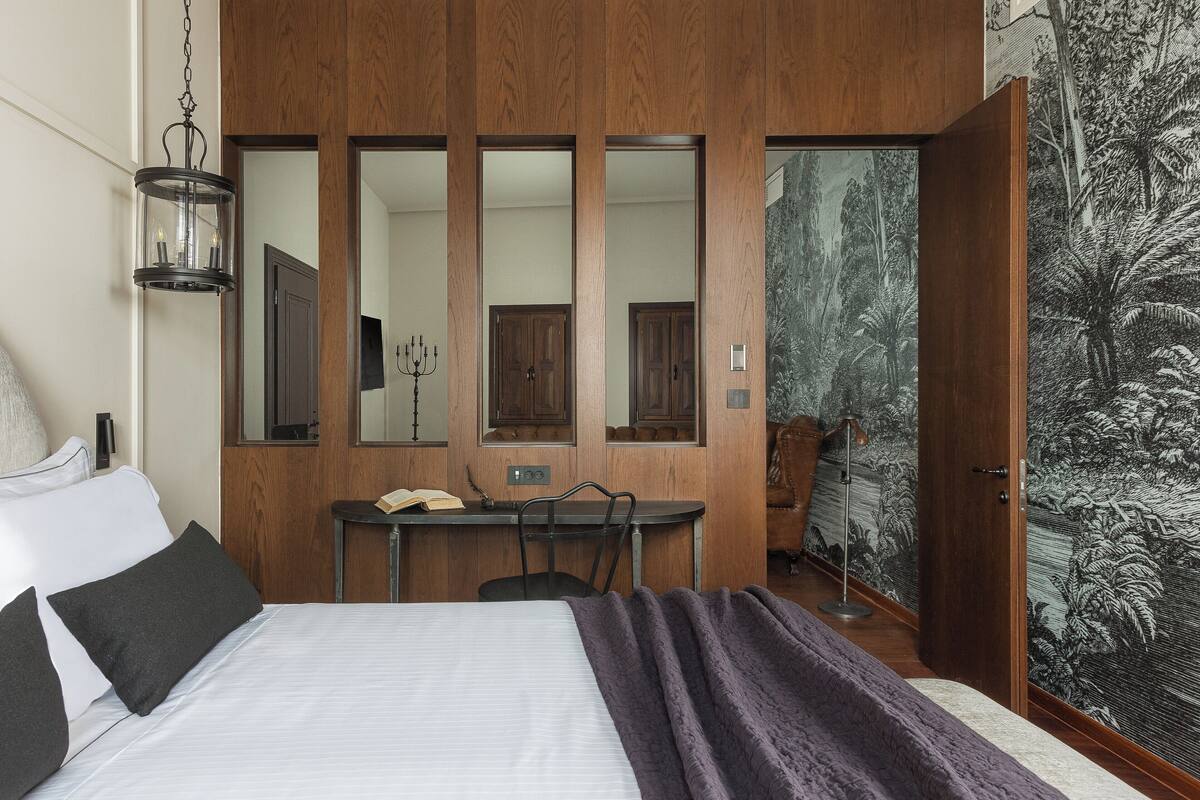 A bedroom with a bed, a desk, a chair, a mirror, and a door leading to another room.