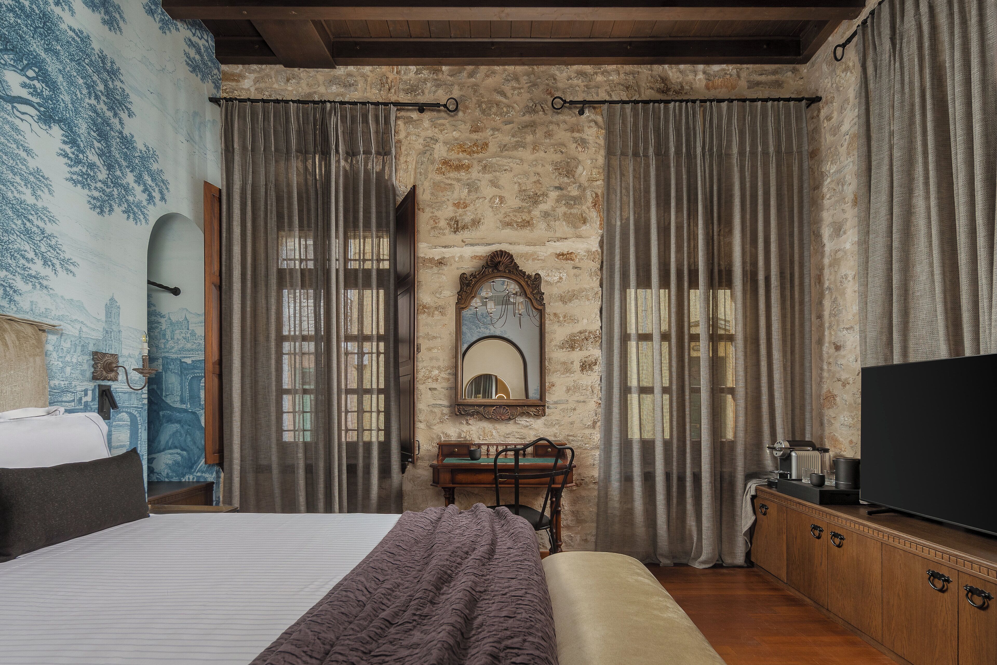 A bedroom with a large bed, a wooden dresser, a television, and a stone wall.