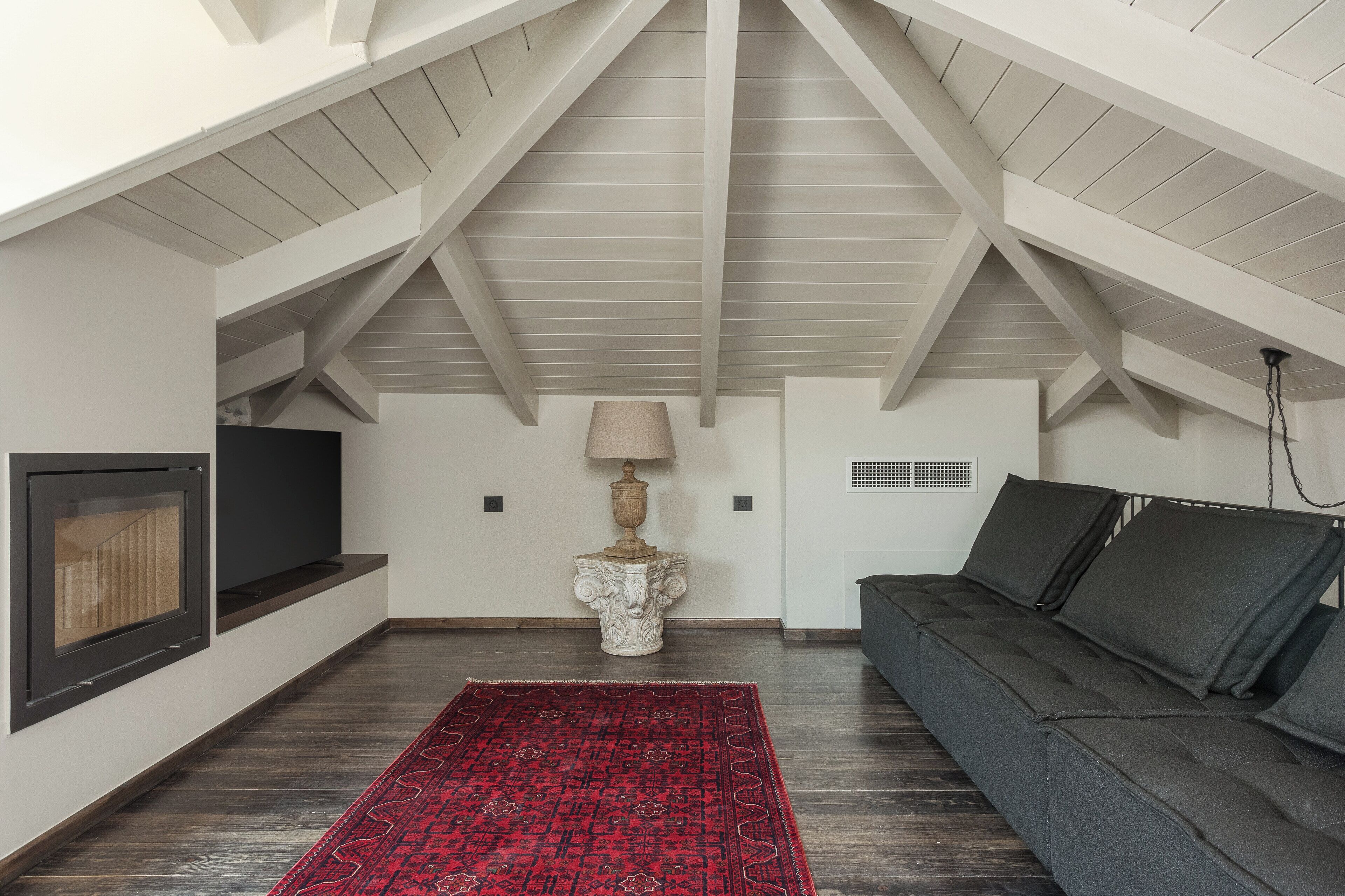 Habitación de ático con piso de madera oscura, alfombra roja estampada, sofá oscuro, lámpara blanca y chimenea.