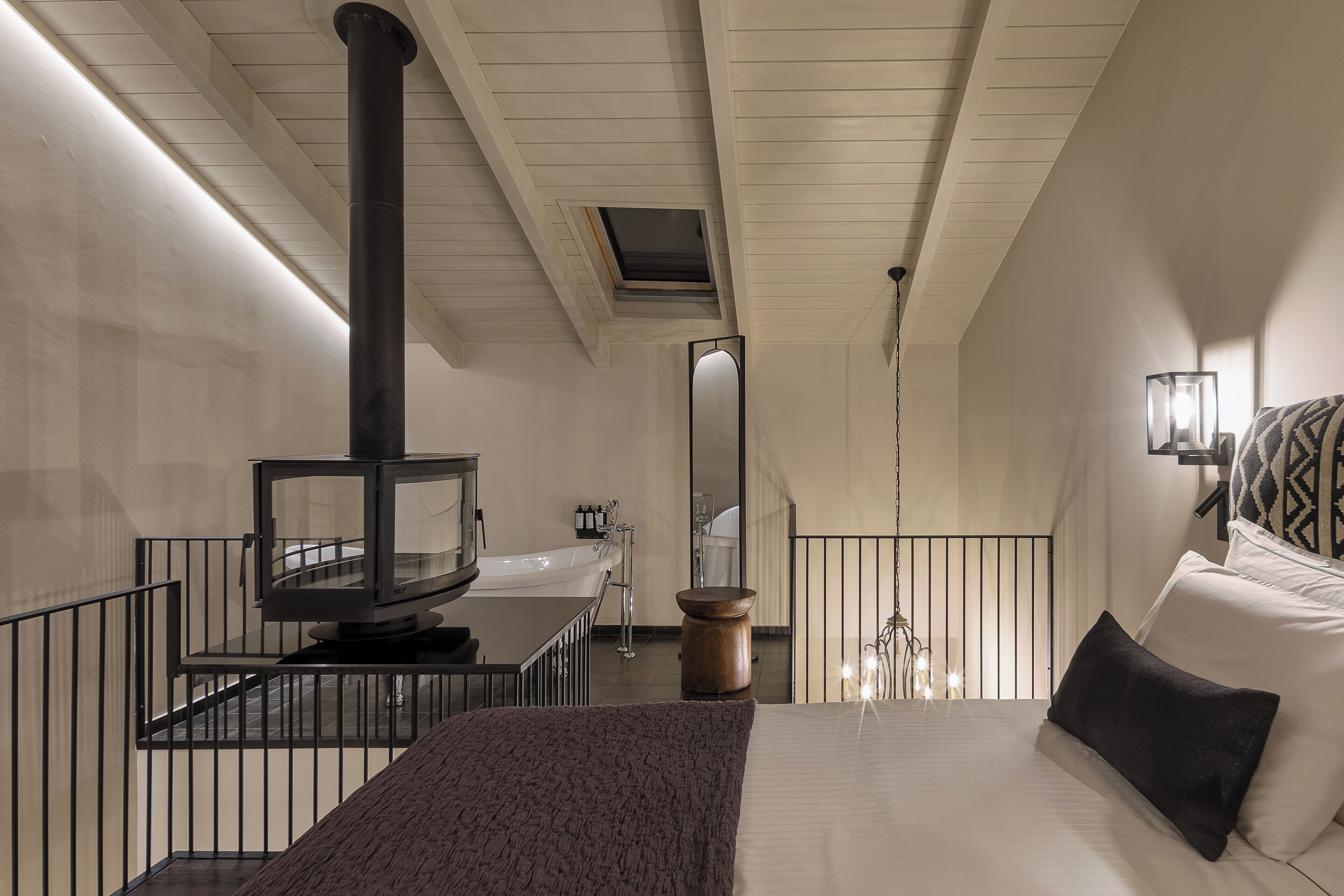 A modern hotel room with a large bed, a wooden floor, a black heater, a bathroom with a bathtub, and a small balcony with a railing.