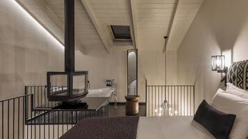 A modern hotel room with a large bed, a wooden floor, a black heater, a bathroom with a bathtub, and a small balcony with a railing.