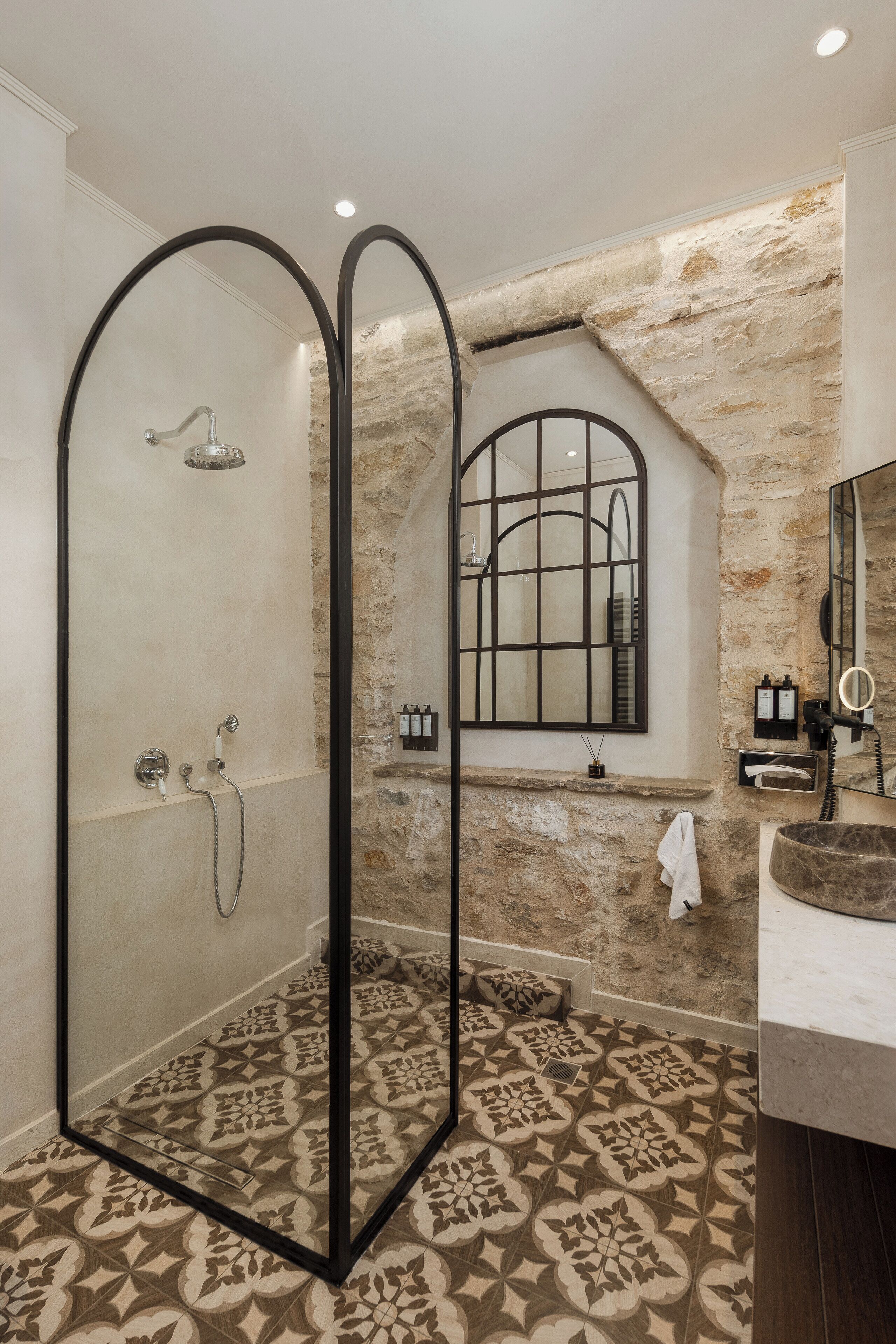 A bathroom with a shower, a sink, and a mirror. The shower has a glass door, and the sink is made of marble. The floor has a patterned tile design.