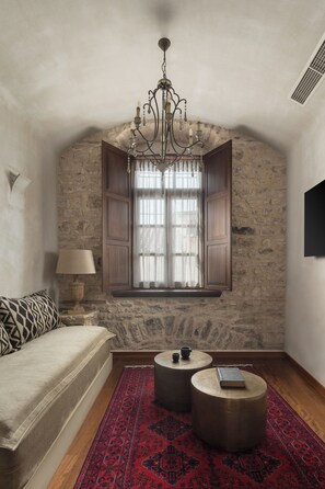 A cozy room with a stone wall, a wooden window, a chandelier, a sofa, two round tables, and a red patterned rug.