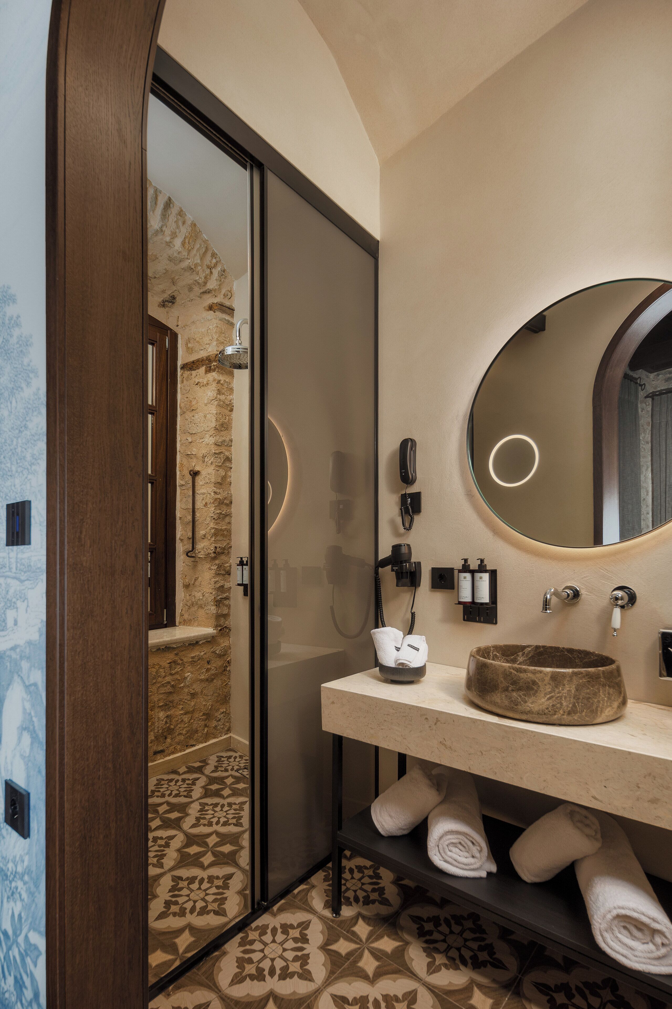 A modern bathroom with a stone wall, a round mirror, a sink with a stone basin, and a glass shower enclosure.
