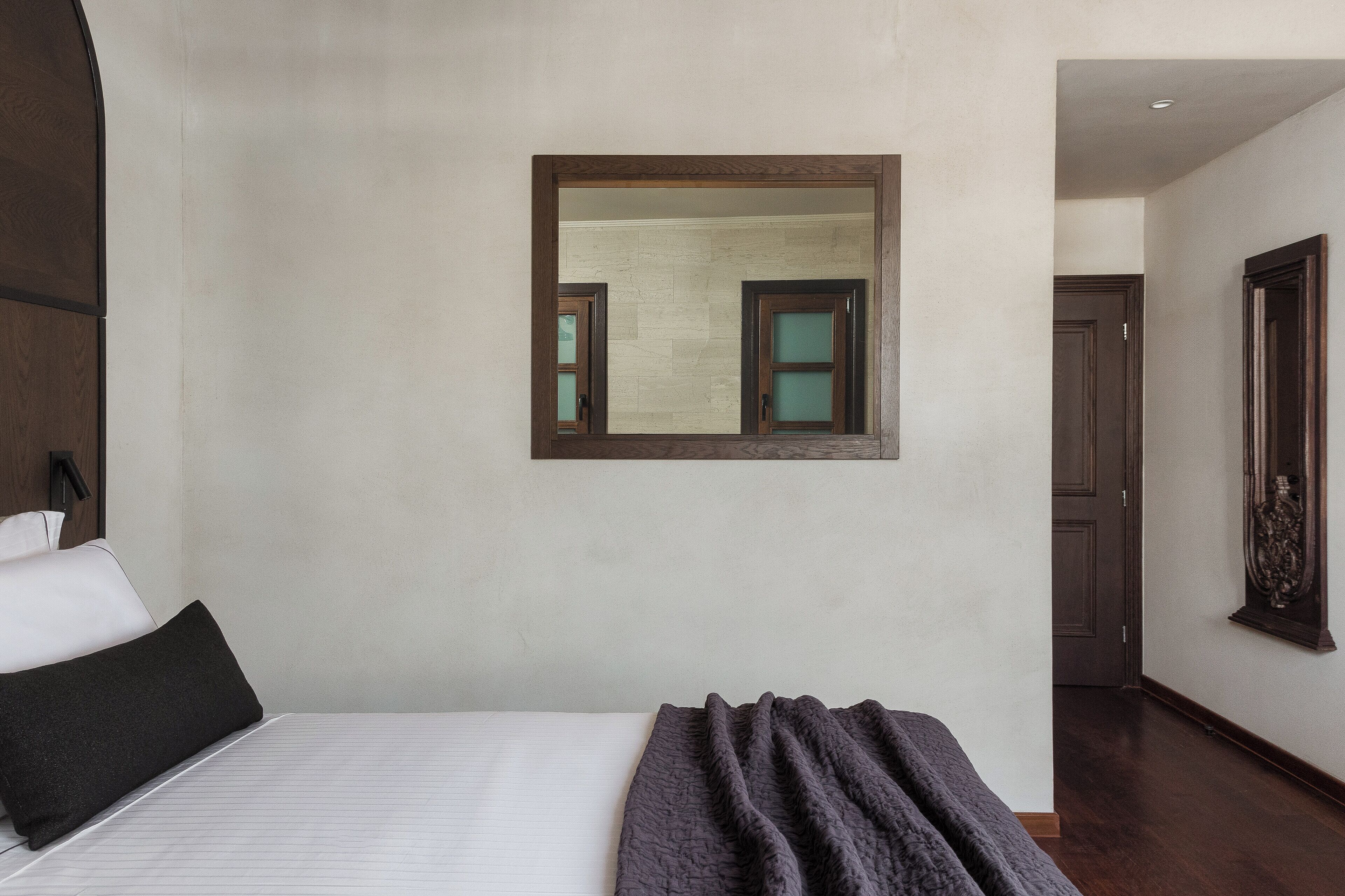 A hotel room with a large bed, a dark-colored blanket, a mirror on the wall, and a wooden door in the background.