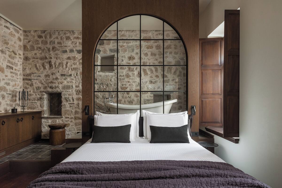 A bedroom with a stone wall, a large arched window, a bed with white linens, a wooden dresser, and a wooden cabinet.
