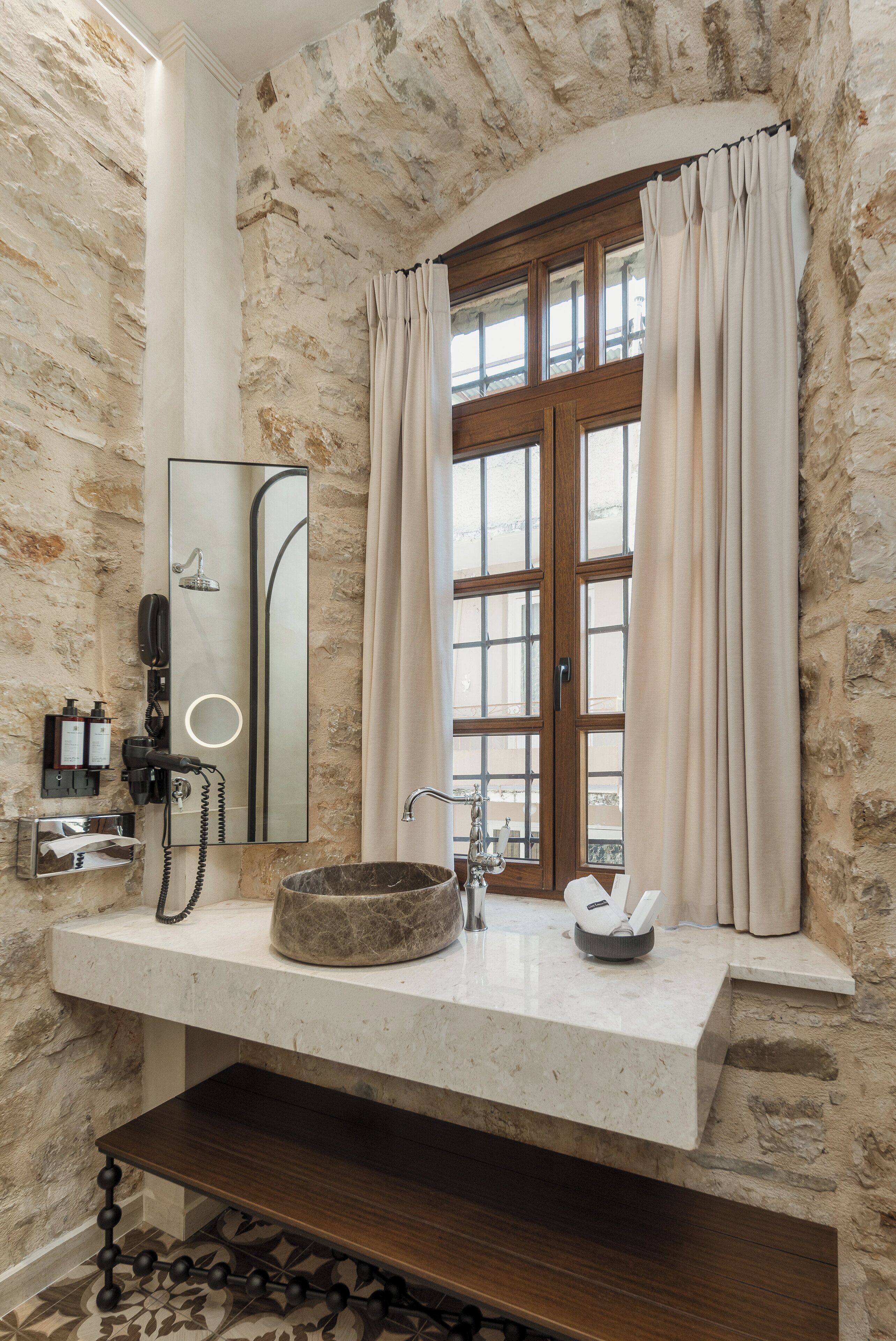 A bathroom with a stone wall, a wooden-framed window with curtains, a sink with a stone basin, a mirror, a hairdryer, and a telephone.