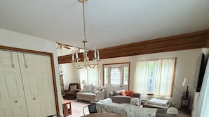 Un comedor con una mesa de madera, sillas negras y una araña. Se ve una cama con colcha y almohadas al fondo.