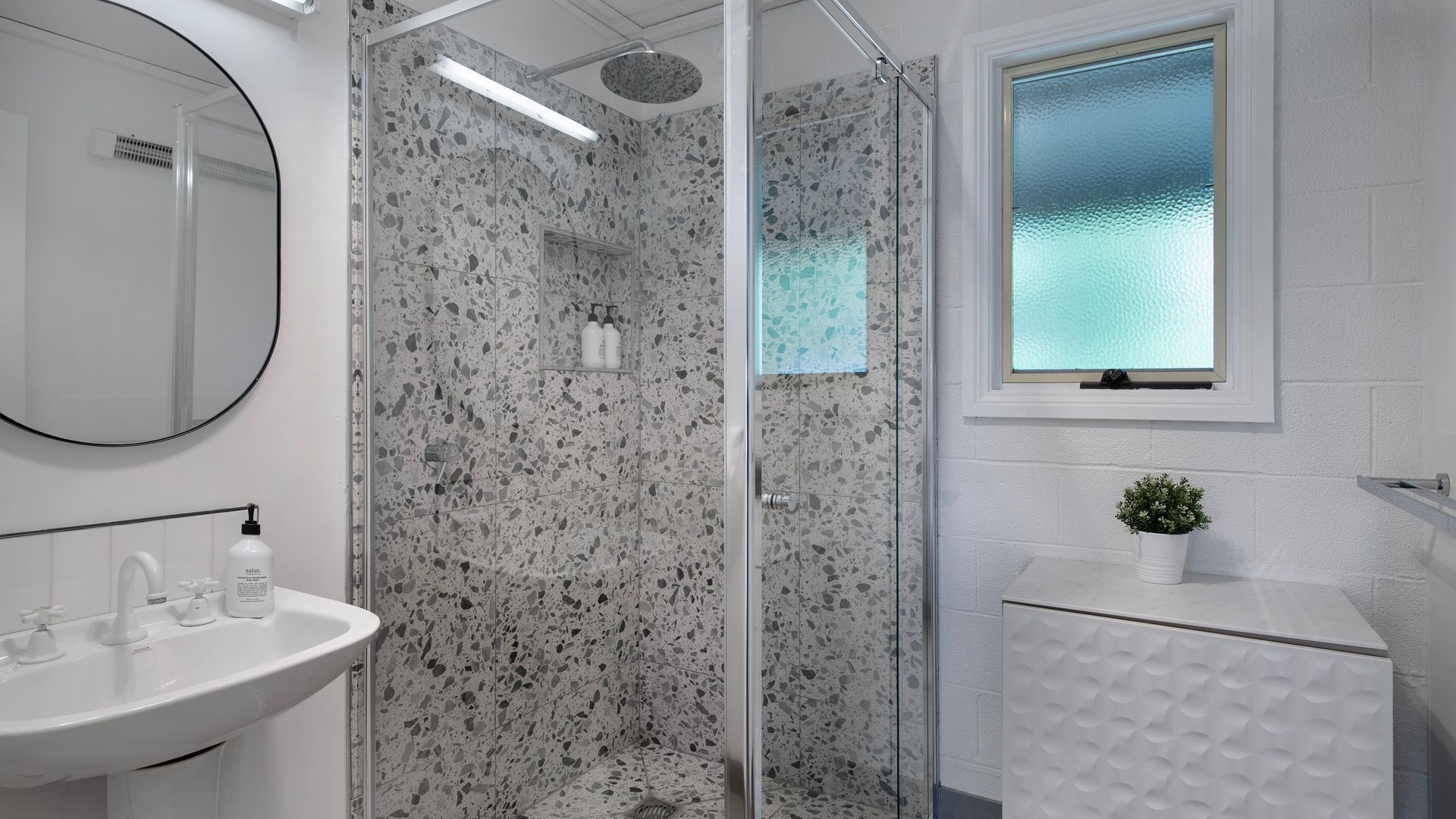 Un baño moderno con cabina de ducha de vidrio, un lavamanos blanco y una ventana con cristal satinado.