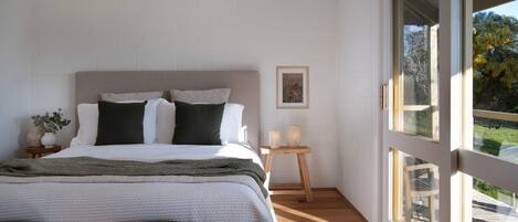 Een slaapkamer met een groot bed, een klein nachtkastje, een ingelijst kunstwerk aan de muur en een raam met uitzicht op bomen.