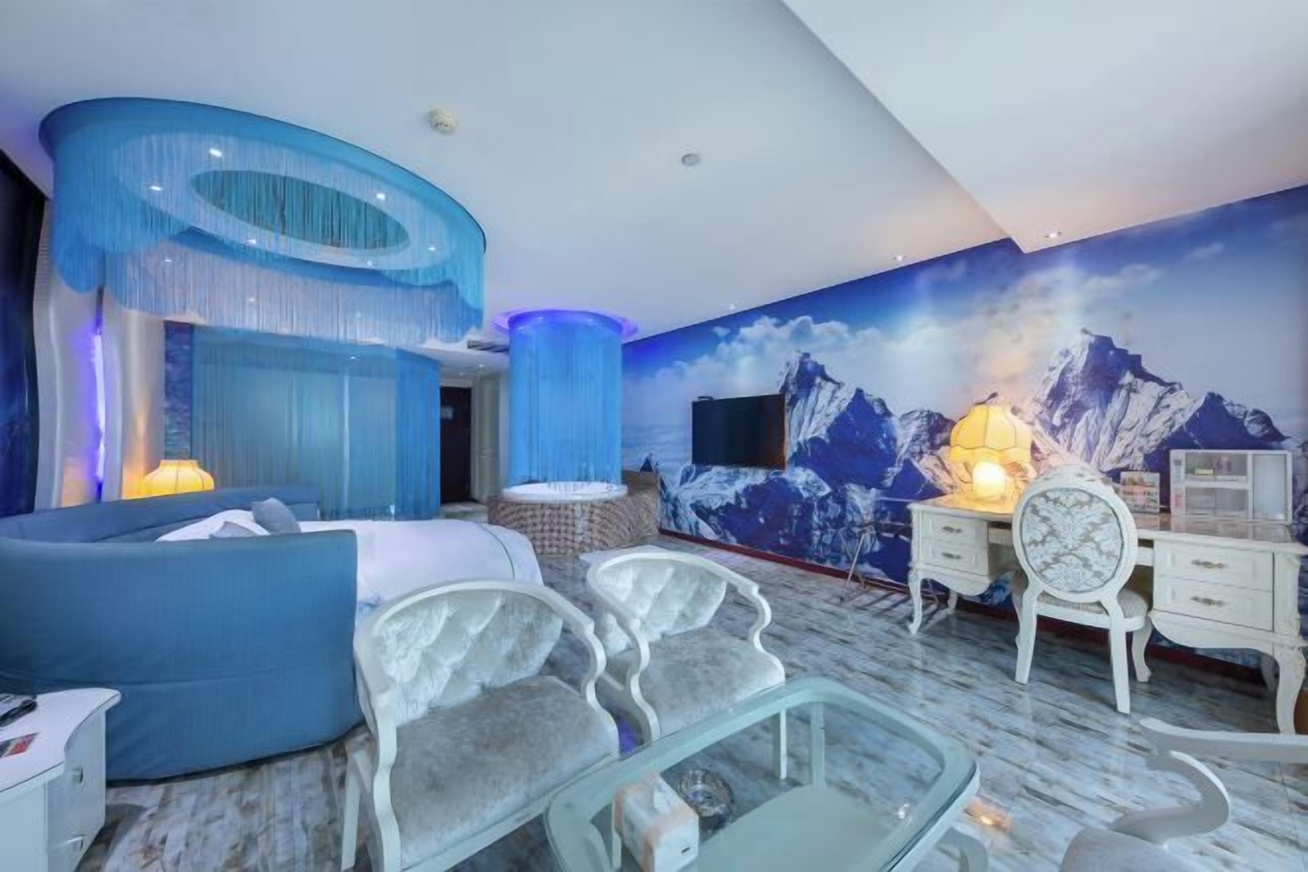 A modern bedroom with a blue accent wall, a mountain mural, a glass coffee table, and a white dresser.