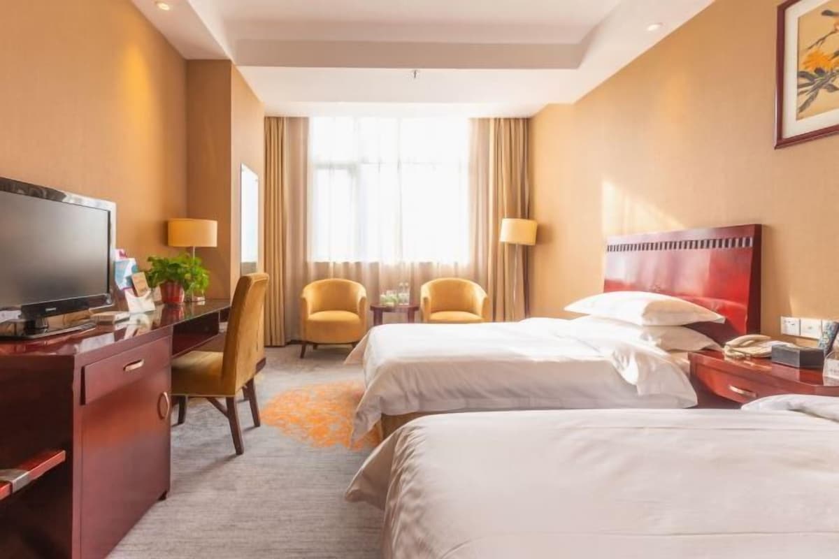Camera d'albergo con due letti, una scrivania con televisore, due poltrone, una finestra con tende e un quadro appeso al muro.