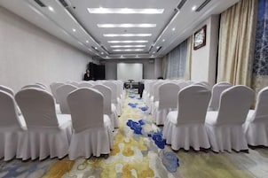 Un salón de conferencias con filas de sillas blancas, una alfombra de estampado floral y una pantalla de proyector al frente.
