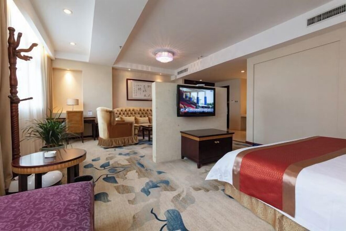 Una camera d'albergo con un letto, una TV, un'area salotto con un divano e poltrone, un tavolino e una pianta in vaso.