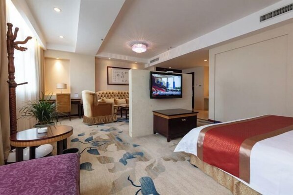 Una camera d'albergo con un letto, una TV, un'area salotto con un divano e poltrone, un tavolino e una pianta in vaso.