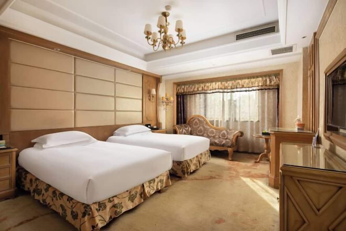 Une chambre d’hôtel avec deux lits, une tête de lit en bois, un lustre, une fenêtre avec des rideaux et un bureau avec un miroir.
