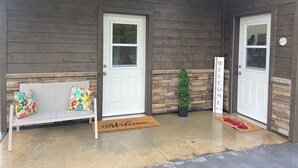 Fachada de madera con dos puertas blancas, un banco con cojines coloridos, un felpudo y una pequeña planta en maceta.