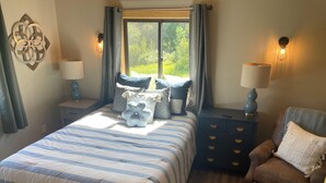 Un dormitorio con cama, mesitas de noche, una silla y una ventana con cortinas.