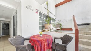 Un comedor con una mesa redonda y sillas de mimbre, un mantel rojo y una planta en maceta con flores amarillas.