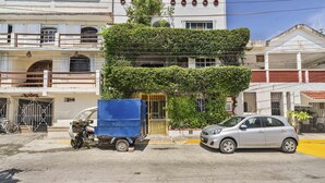 Un edificio con una fachada verde y original cubierta de hiedra, una camioneta azul estacionada frente a él y un coche plateado en la calle.