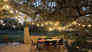 Un patio exterior con chimenea de piedra, mesa y sillas de madera, y luces colgantes.