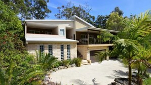 Una casa moderna de dos pisos con balcón, rodeada de abundante follaje y un jardín cuidado.
