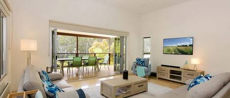 A spacious living room with a sofa, coffee table, and a TV mounted on the wall. There is a balcony with a table and chairs, and a sliding glass door leading outside.