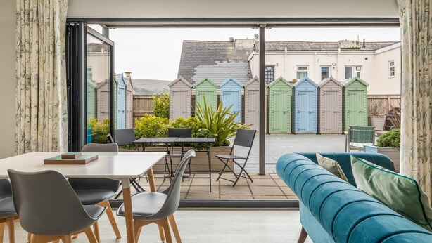 A modern living room with a sofa, dining table, chairs, and a view of colorful sheds in the background. - 2 Bed in Woolacombe (oc-b27615) (Woolacombe)